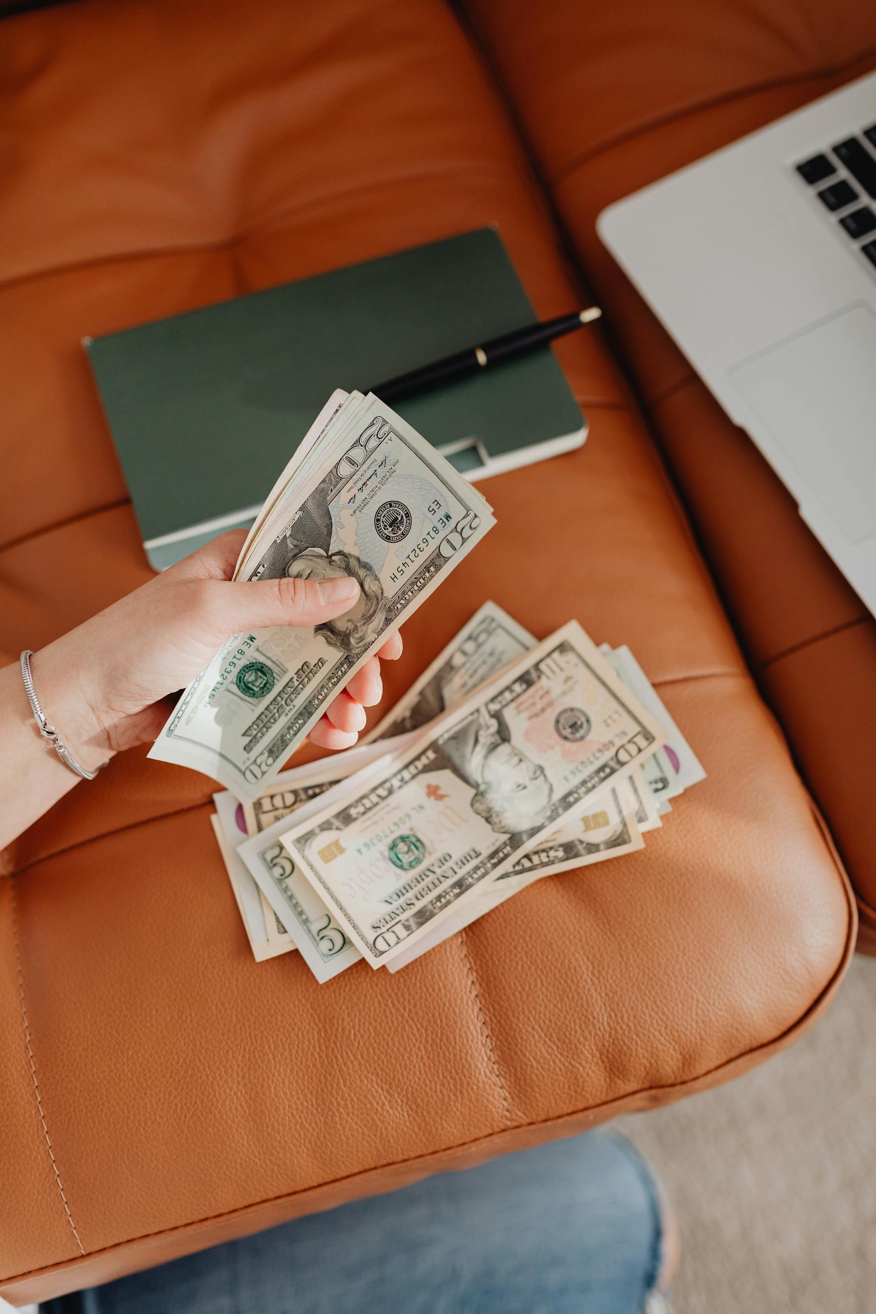 Person counting US dollars on a leather couch with a notebook and laptop nearby.