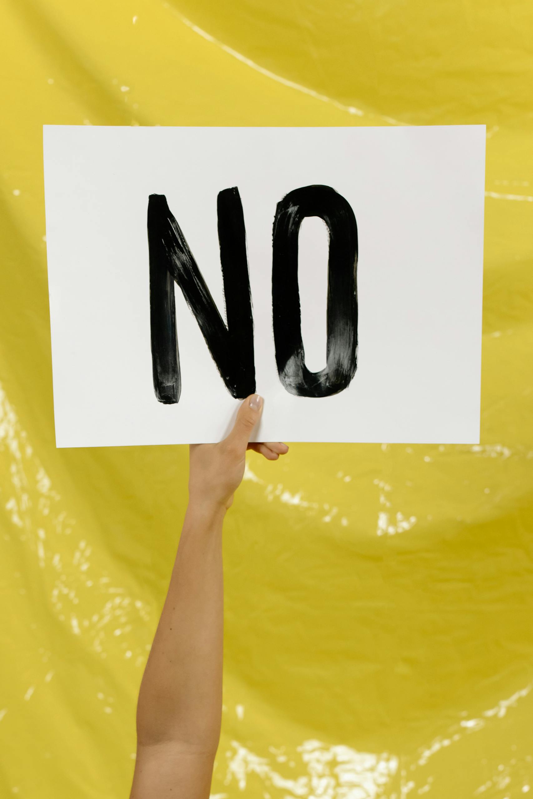 A hand holding a 'NO' sign against a bright yellow background.