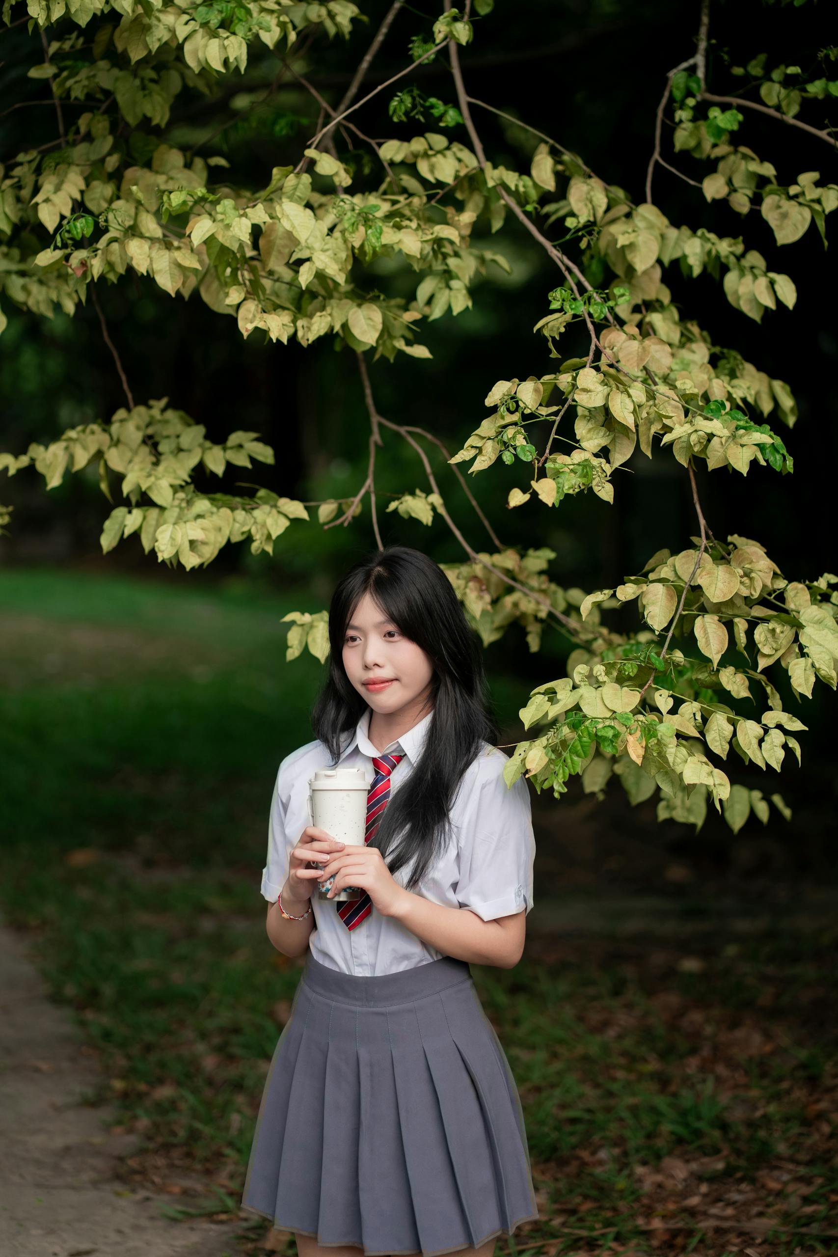 Young woman in school uniform holding a beverage under a leafy tree canopy outdoors.