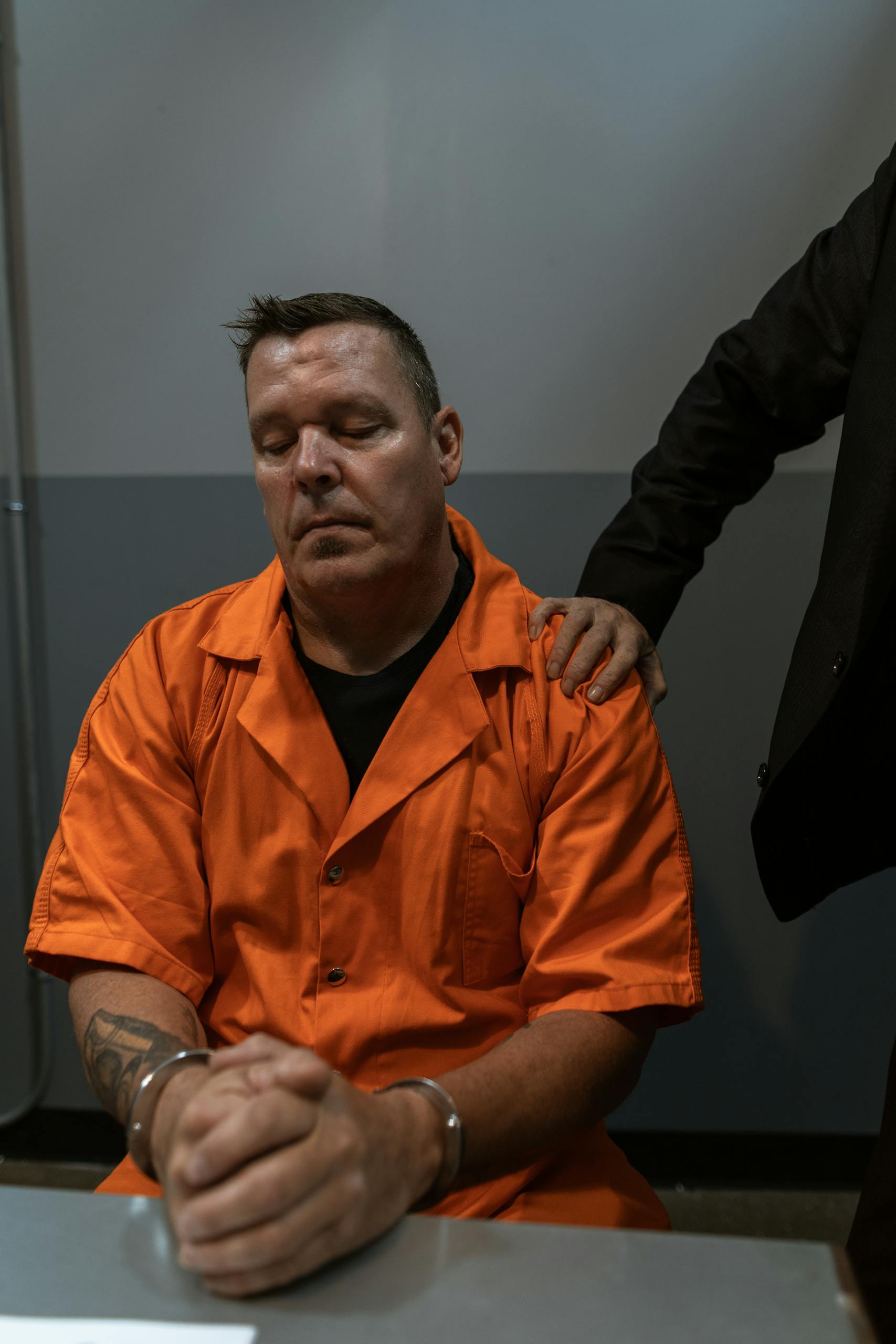 A prisoner in an orange jumpsuit sits handcuffed under supervision indoors.