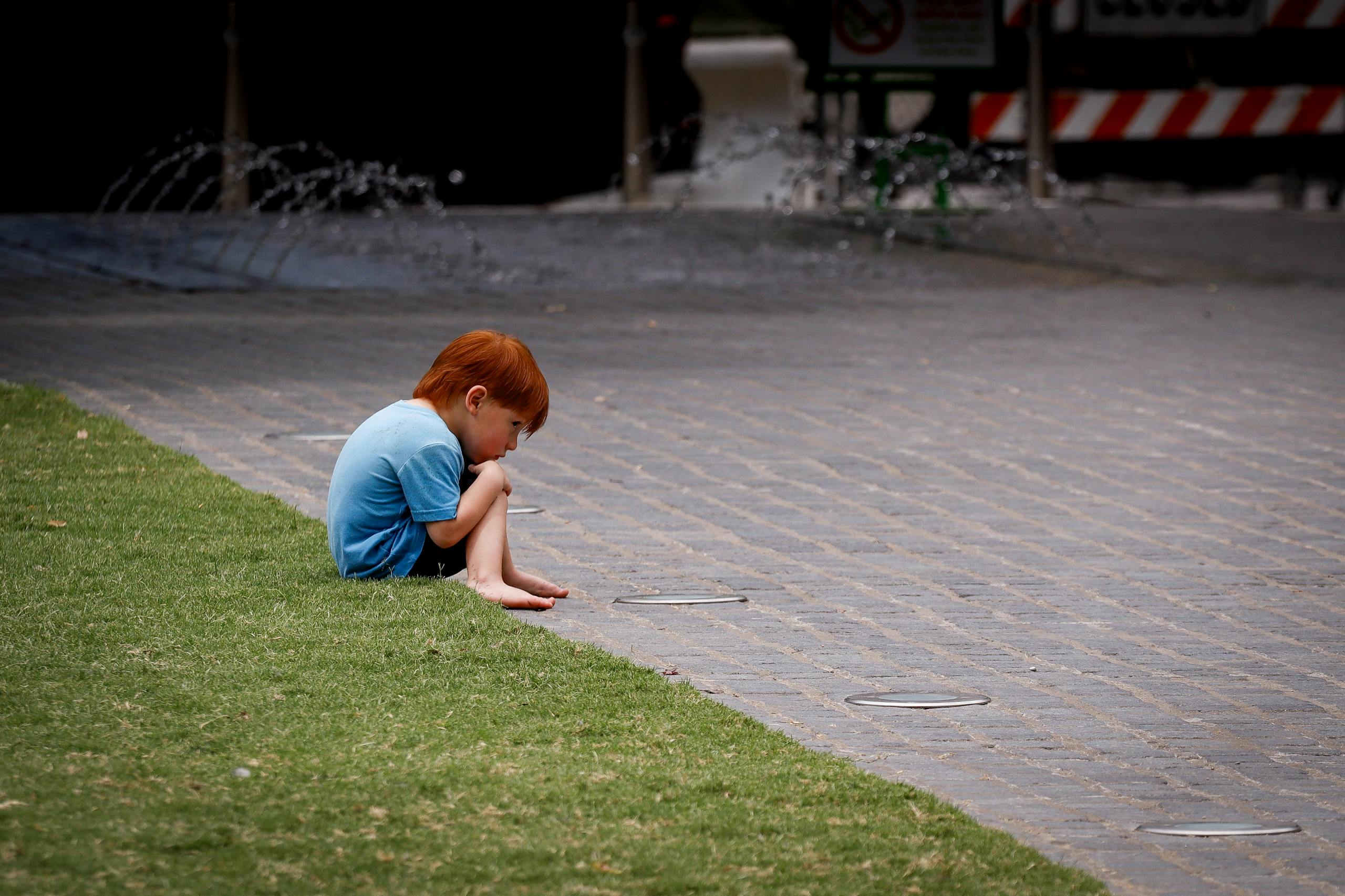 A young red-haired child sits thoughtfully on the pavement near a grassy area in an urban setting.